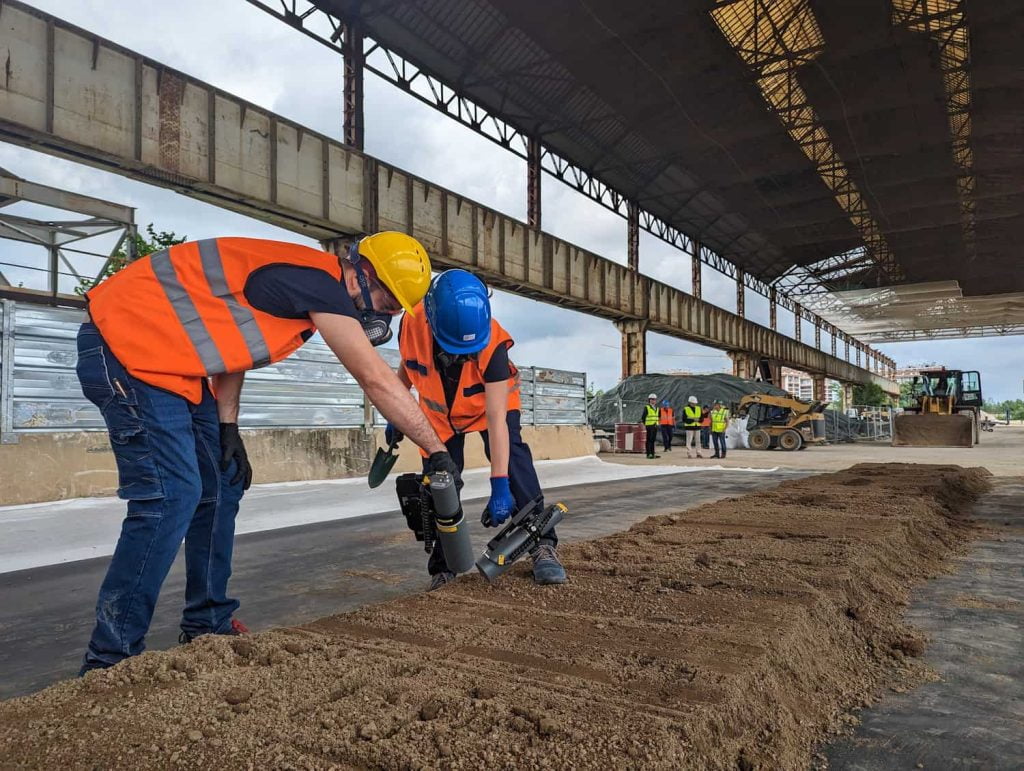 due operatori che lavorano nella radiografia industriale con strumenti specifici sul terreno. Sono all'interno di un complesso di lavoro con scavatori e terra da analizzare.