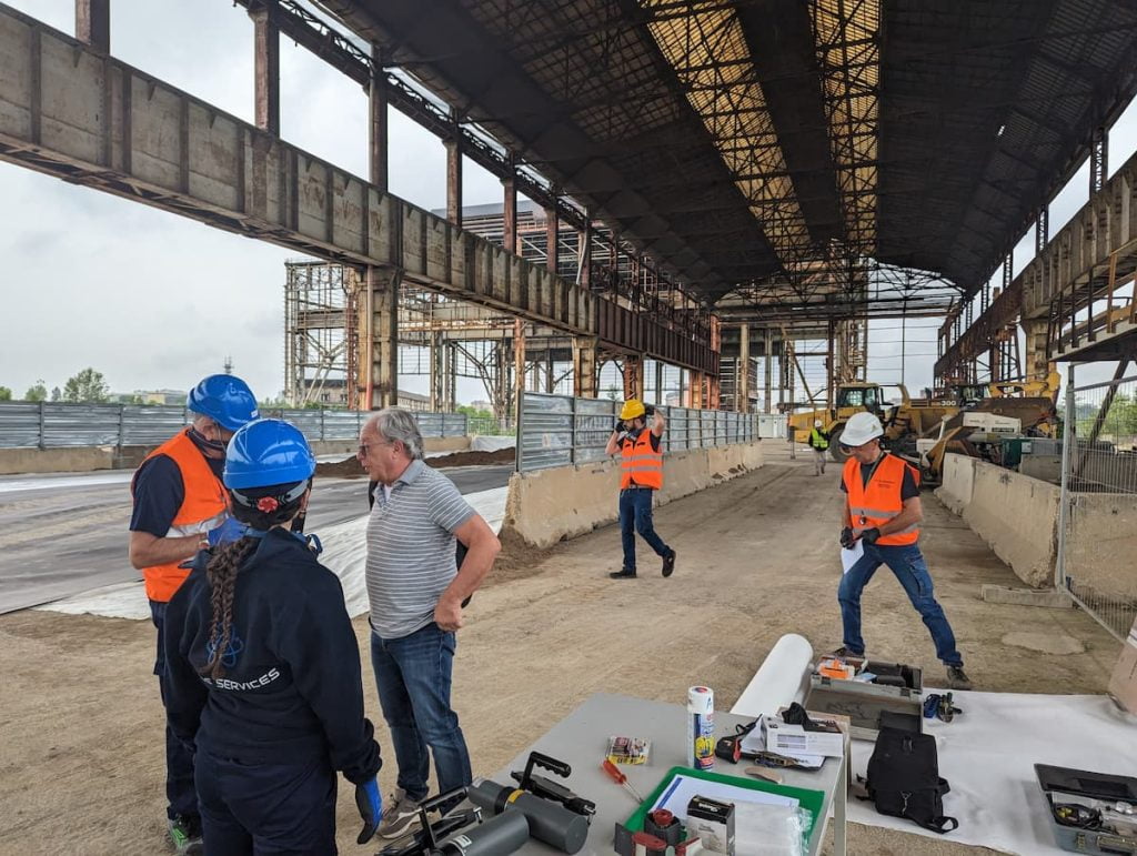 team Atlas Services competente in radiografia per saldatura. Cinque lavoratori del team che discutono sul da farsi. Nello sfondo scavatori e oggetti di lavoro nel cantiere.